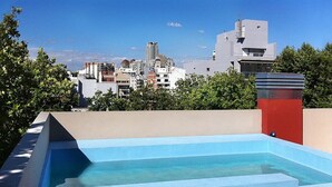 A rooftop swimming pool with a wooden deck, surrounded by trees and buildings in the background.