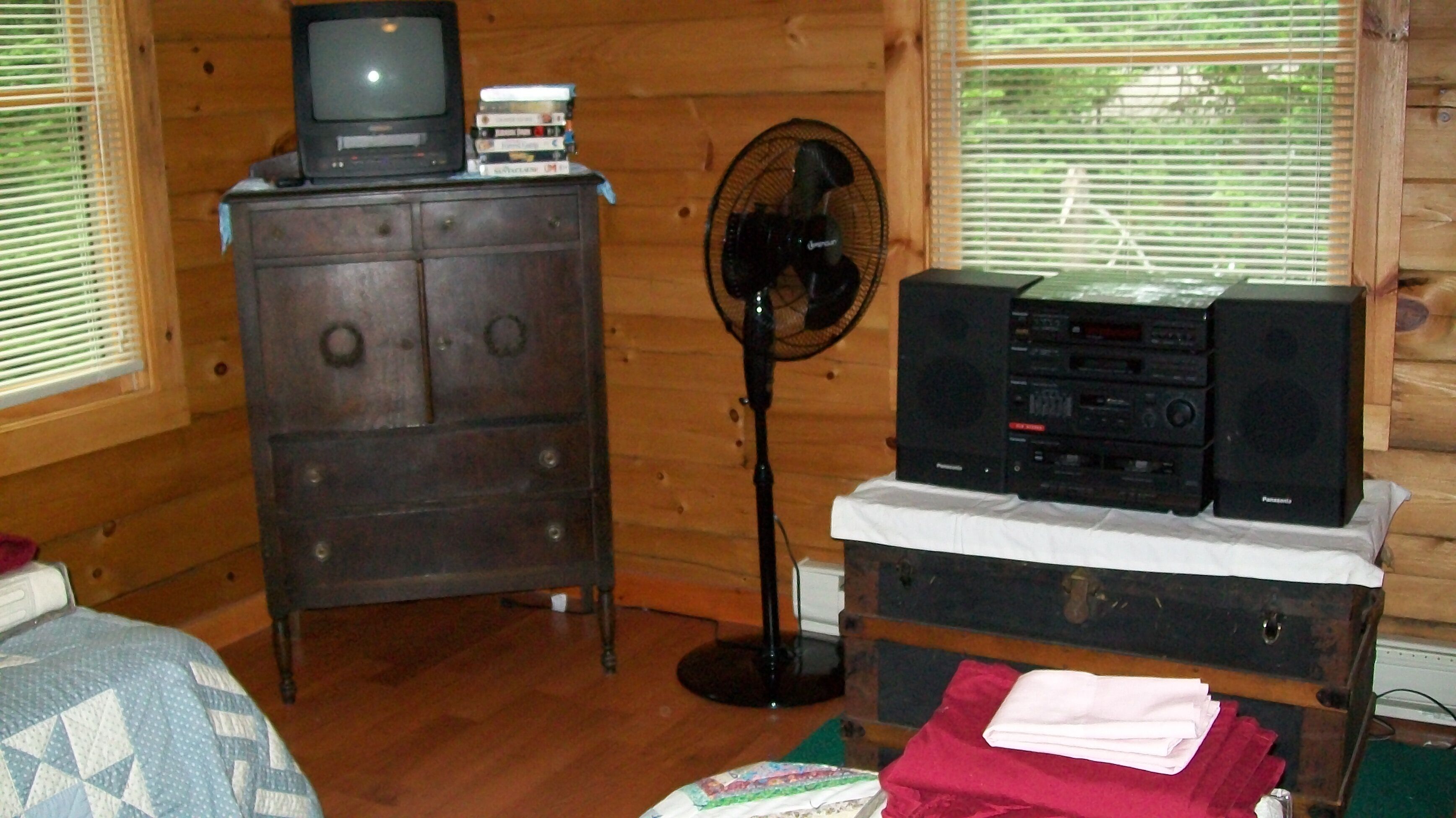 A wooden cabin room with a wooden dresser, a television, a stack of DVDs, a fan, and a stereo system.
