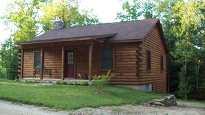 Une cabane en rondins avec un porche à l’avant, une cheminée et un petit jardin devant.