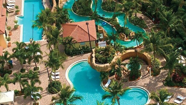 An aerial view of a resort with multiple swimming pools, palm trees, and a small building with a red roof. - Bay Bungalow at Naples Bay Resort (Naples)