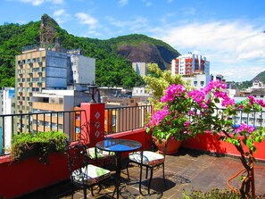 Eine Dachterrasse mit Tisch und Stühlen, einem Blumentopf mit rosa Blüten und Blick auf eine Stadtlandschaft mit Gebäuden und einem Berg.