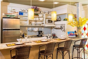 A modern kitchen with a central island, stainless steel appliances, wicker chairs, and pendant lighting.