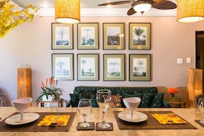 A dining area with a table set for two, framed tropical landscape pictures on the wall, a green sofa, and a ceiling fan.