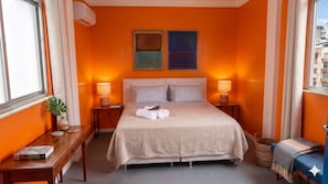 A hotel room with a bed, bedside tables, a nightstand, a window, and a view of buildings outside.