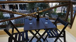 A table with two chairs, a bottle, and two glasses on a balcony with a chain-link fence and a view of buildings and trees.