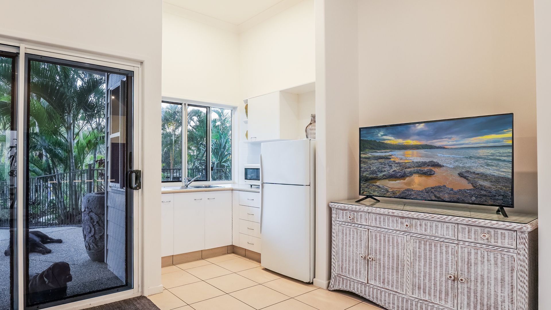 A modern kitchen with a large TV, white cabinets, and a view of a tropical outdoor area.