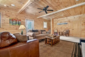 A cozy wooden cabin interior with a leather sofa, a wooden coffee table, and a ceiling fan. - Meadow Creek - Pet-friendly retreat with rambling creek, hot tub, fire-pit, WiFi, and sprawling manicured meadow land for serene relaxation (Morganton)