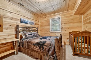 Une chambre à coucher dans un chalet en bois, avec un lit, un berceau, une fenêtre avec des stores et un tableau accroché au mur.