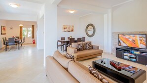 Een ruime woonkamer met een bank, salontafel en tv-meubel. Er is een eethoek met een tafel en stoelen, en een keuken is zichtbaar op de achtergrond.