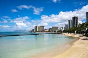 Ein Strand mit klarem türkisblauem Wasser, einem sandigen Ufer und hohen Gebäuden im Hintergrund.