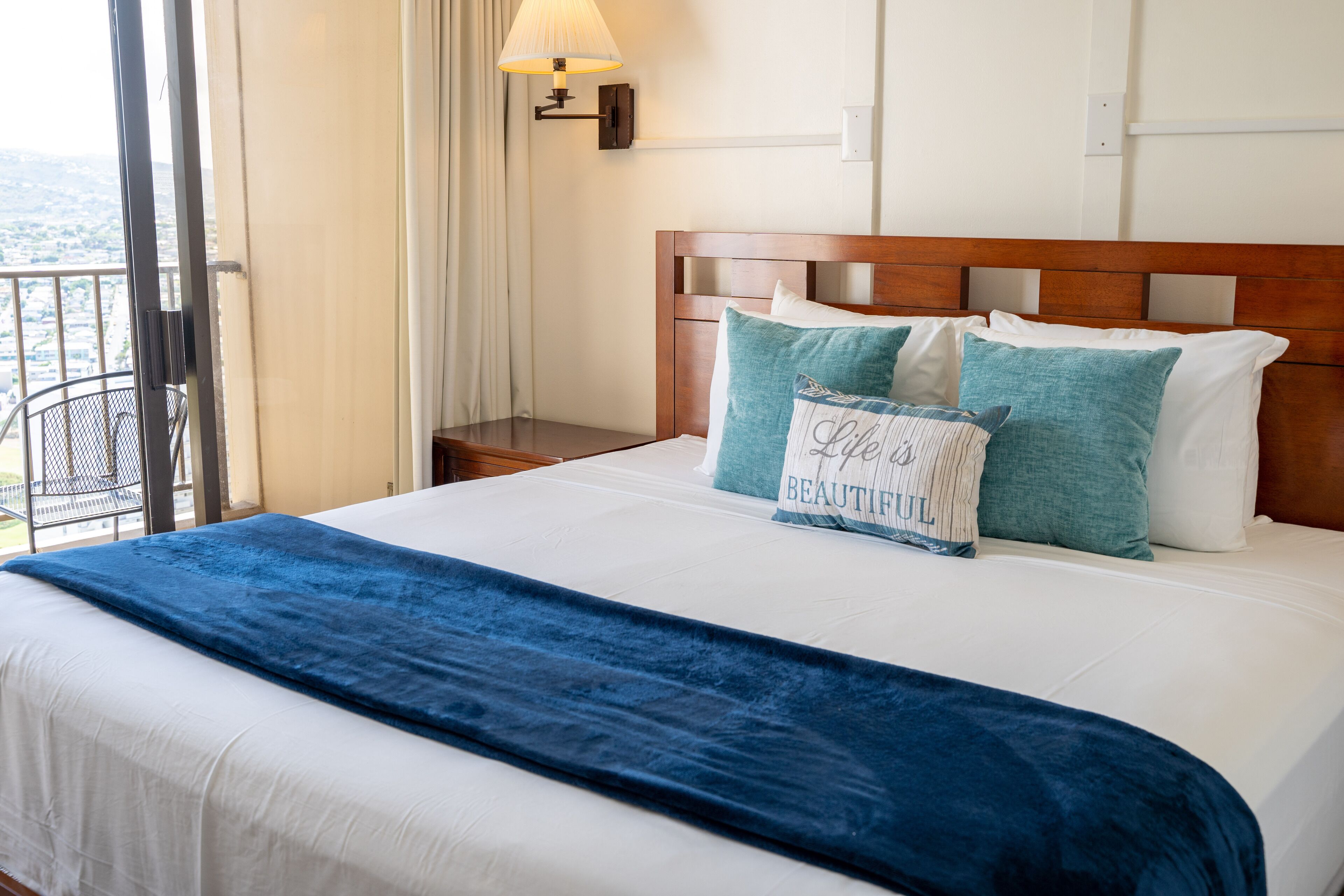A hotel room with a bed, a nightstand, a lamp, and a view of the city.