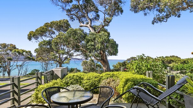 A patio with outdoor seating, a view of the sea, and a large tree. - Apartment 2 Kalimna (Lorne)
