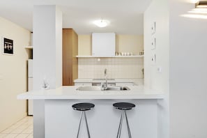 A modern kitchen with a white countertop, stainless steel sink, and two black stools. - Apartment 2 Kalimna (Lorne)