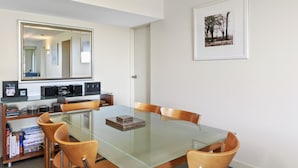 A dining area with a glass table, wooden chairs, a bookshelf with books and audio equipment, a large mirror, and a framed picture on the wall. - Apartment 2 Kalimna (Lorne)