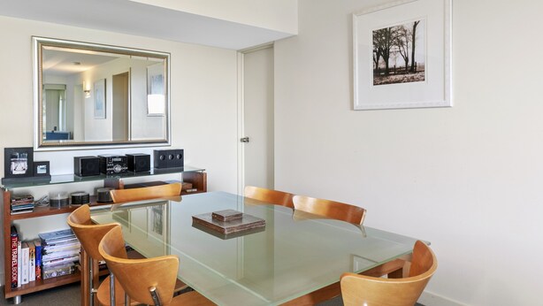 Een eethoek met een glazen tafel, houten stoelen, een boekenkast met boeken en audioapparatuur, een grote spiegel en een ingelijste foto aan de muur.