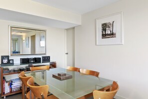 A dining area with a glass table, wooden chairs, a bookshelf with books and audio equipment, a large mirror, and a framed picture on the wall.