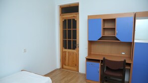 Una habitació amb una porta de fusta, un escriptori amb calaixos, una cadira i un llit.