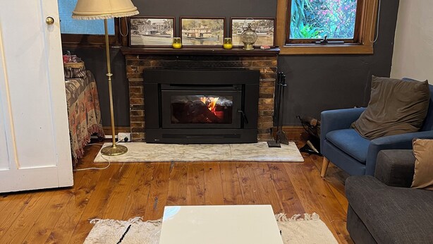 A living room with a fireplace, a blue sofa, a white coffee table, and a window with a view of greenery. - The Grove Gunbower.. (Gunbower)