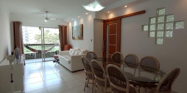 A modern living room with a glass dining table, wicker chairs, a white sofa, and a large window with a view of a building and trees.