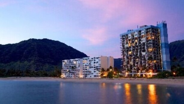 A coastal hotel with multiple buildings, including a tall structure with balconies, situated near a body of water with a mountainous backdrop.