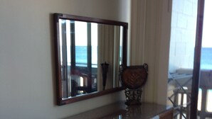A room with a wooden desk, a chair, a large mirror, and a view of the ocean through a sliding glass door.