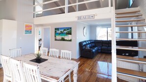A spacious living area with a dining table, white chairs, a black leather sofa, and a staircase leading to an upper level. - Pet-Friendly | 200m to Beach | Sleeps Large Groups | Beach Views | Air Con (Callala Beach)