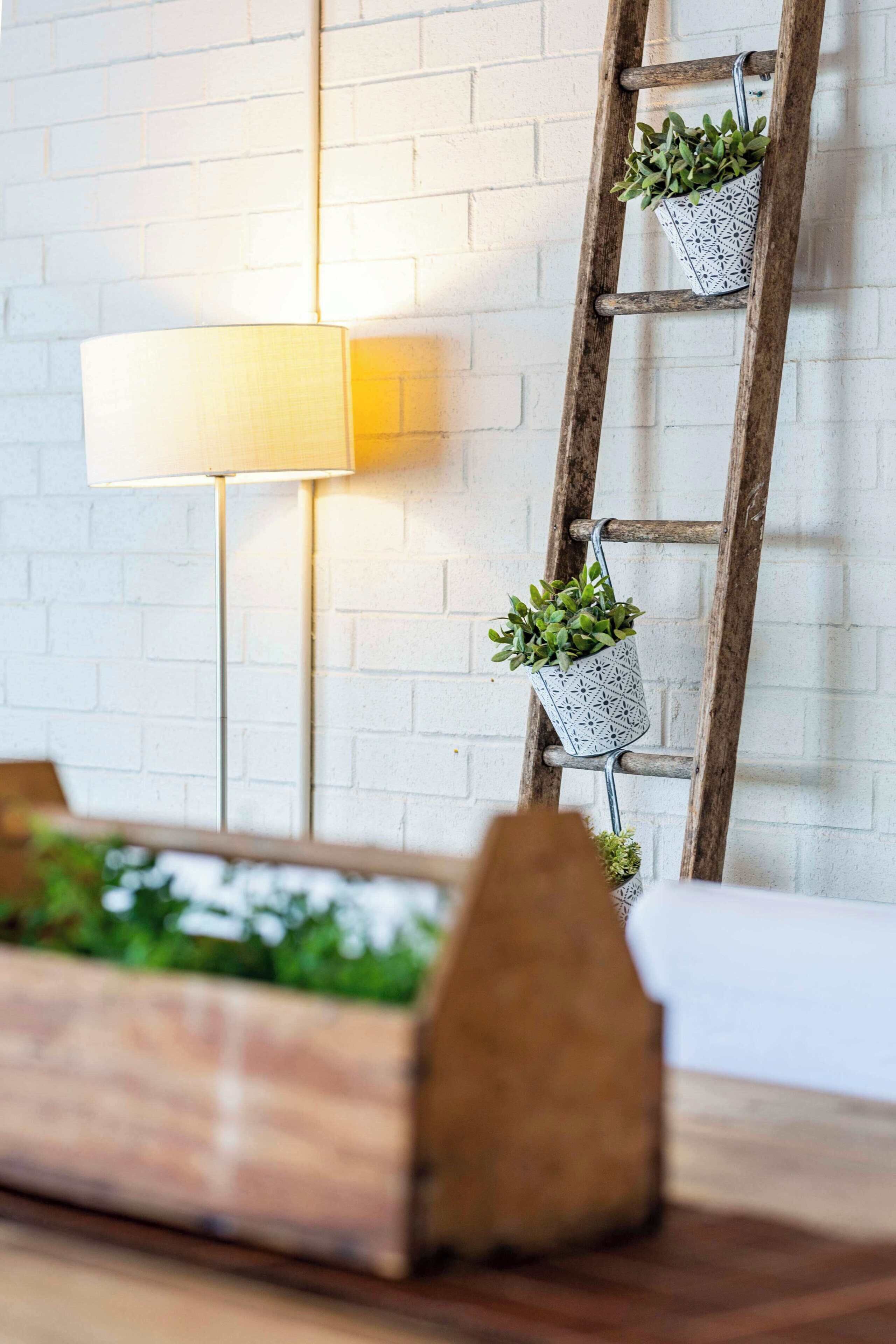 Una jardinera de madera con follaje, una lámpara y una escalera con plantas en macetas frente a un muro de ladrillo blanco.