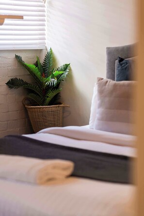 A bed with white bedding, a grey pillow, a potted plant, and a window with blinds. - The Edward Townhouse 155 | Steps to Town (Orange)