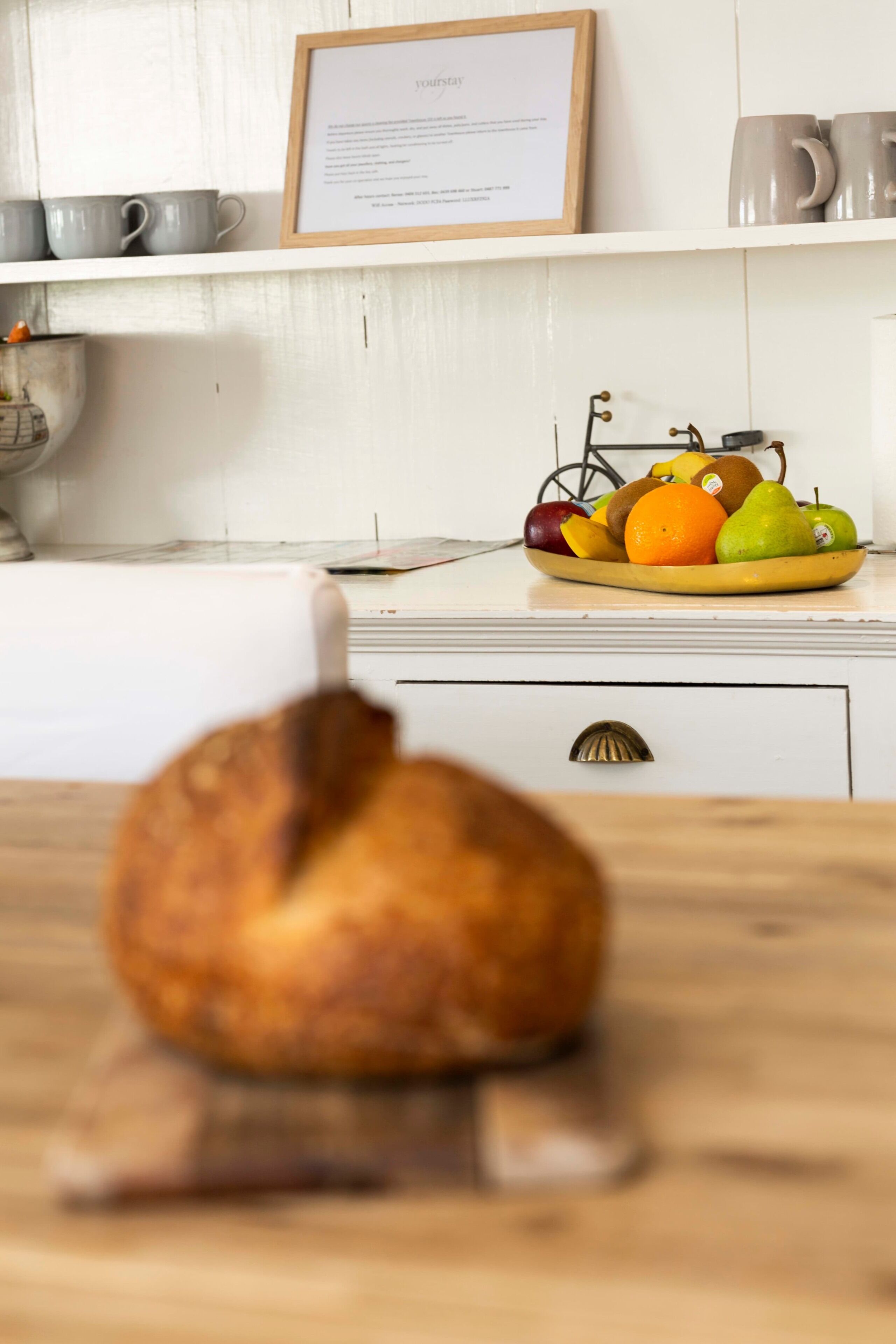 A kitchen with a loaf of bread on a wooden stand, a fruit bowl with apples, oranges, and pears on the counter, and a framed menu on the wall.