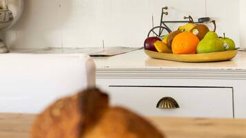 A kitchen with a loaf of bread on a wooden stand, a fruit bowl with apples, oranges, and pears on the counter, and a framed menu on the wall.