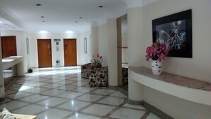 A hotel lobby with a tiled floor, a reception desk, a wall-mounted painting, and a potted plant. - Mi's Apartment - 2 minutes from Pitangueiras Beach - Guarujá (Guarujá)