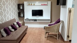 A living room with a brown sofa, a wicker chair, a flat-screen TV on a white wall, and patterned wallpaper. - Mi's Apartment - 2 minutes from Pitangueiras Beach - Guarujá (Guarujá)