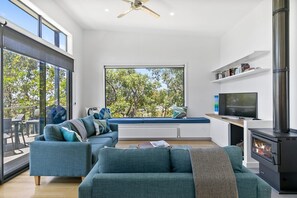 A modern living room with a sofa set, a television, a fireplace, and a large window with a view of trees.