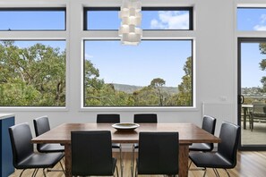 A dining area with a large table and chairs, large windows with a view of trees and hills, and a hanging light fixture.