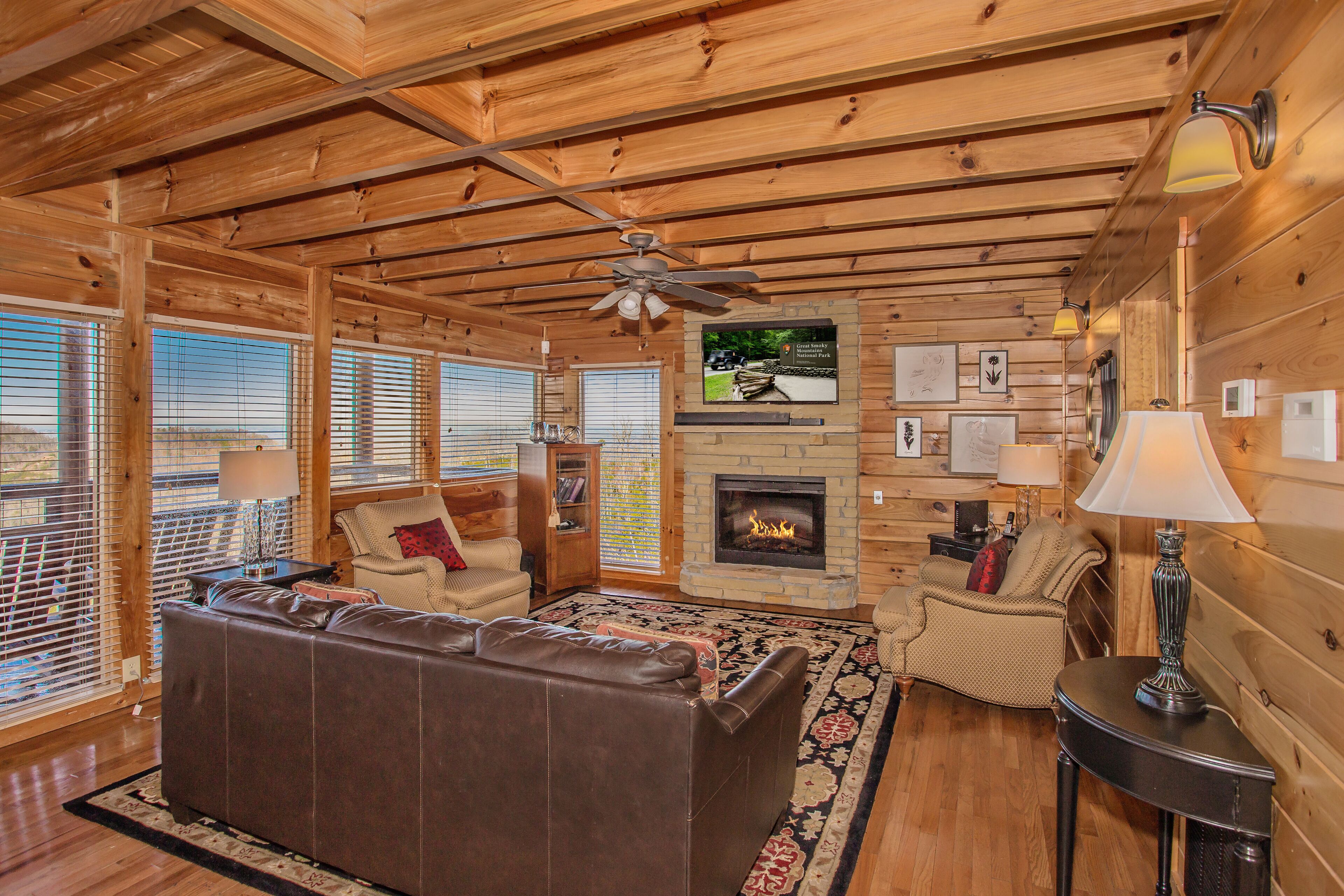 A cozy cabin interior with a fireplace, wooden walls, a leather sofa, and a ceiling fan.