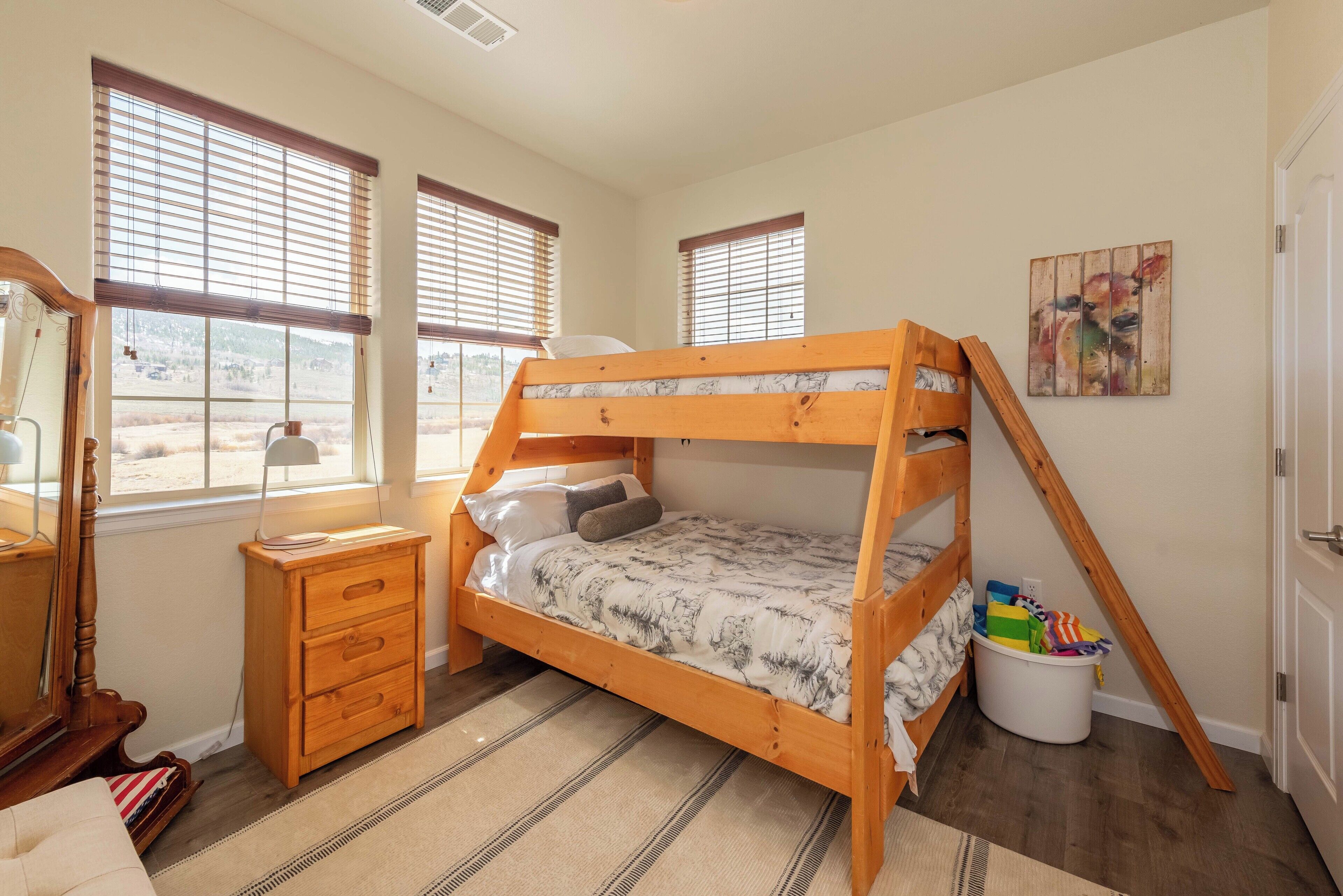 A bunk bed with a wooden frame and a patterned bedspread, a wooden dresser with drawers, a mirror, and a window with blinds.