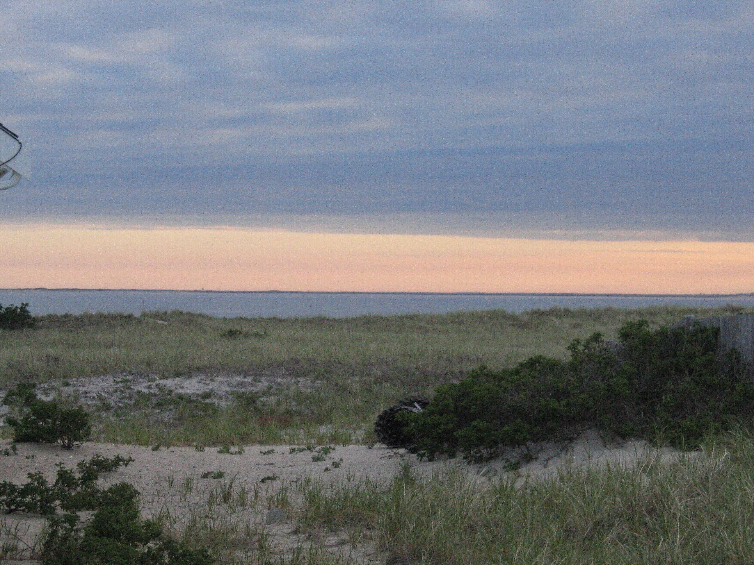 A coastal landscape with a sandy beach, grassy dunes, and a distant shoreline under a cloudy sky.