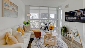 Un salon moderne avec un canapé blanc, une table basse agrémentée d’un vase, un téléviseur fixé au mur et une grande fenêtre donnant sur la ville.