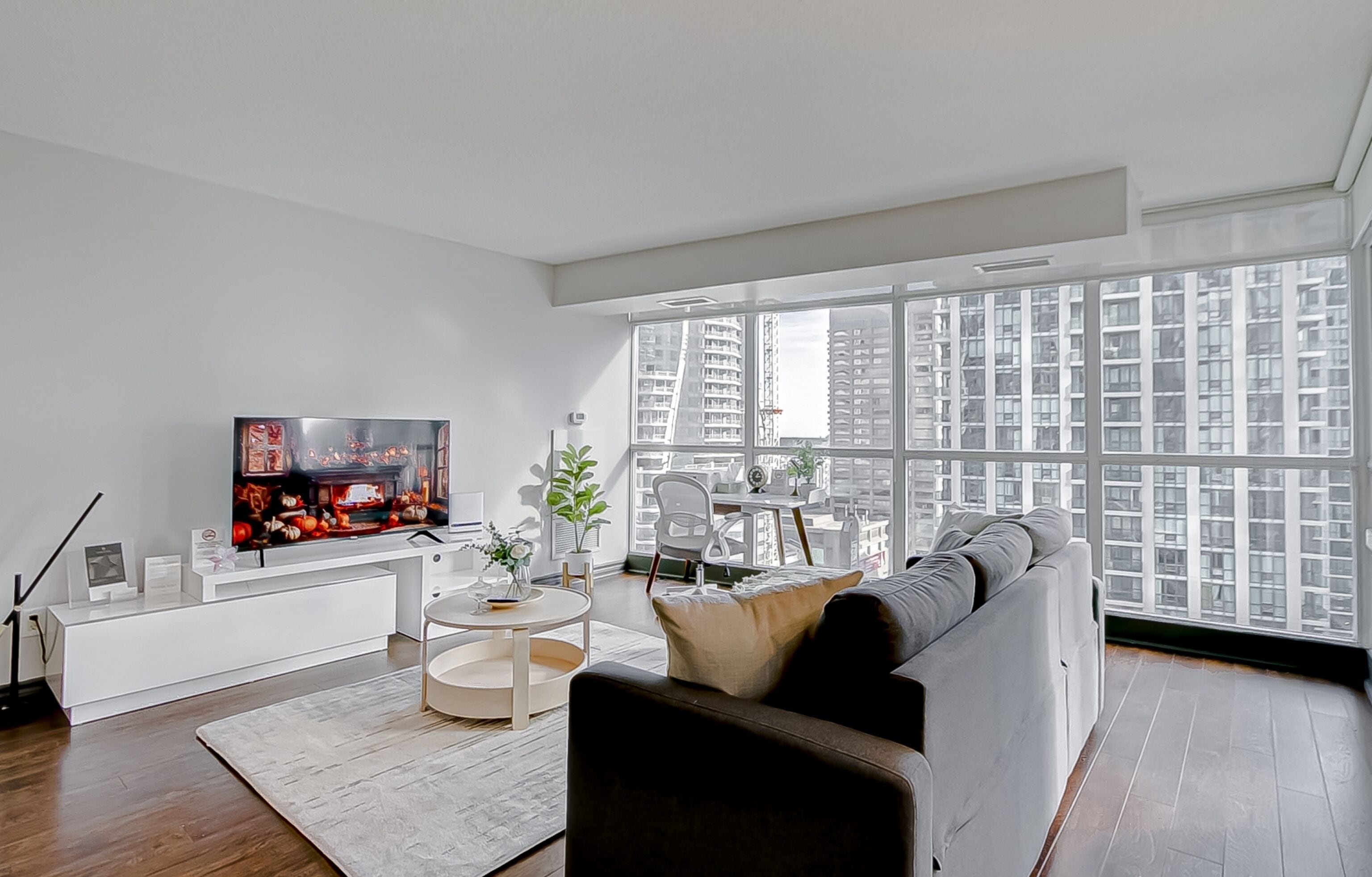 A modern living room with a large TV, a sofa, a dining table, and a view of high-rise buildings.
