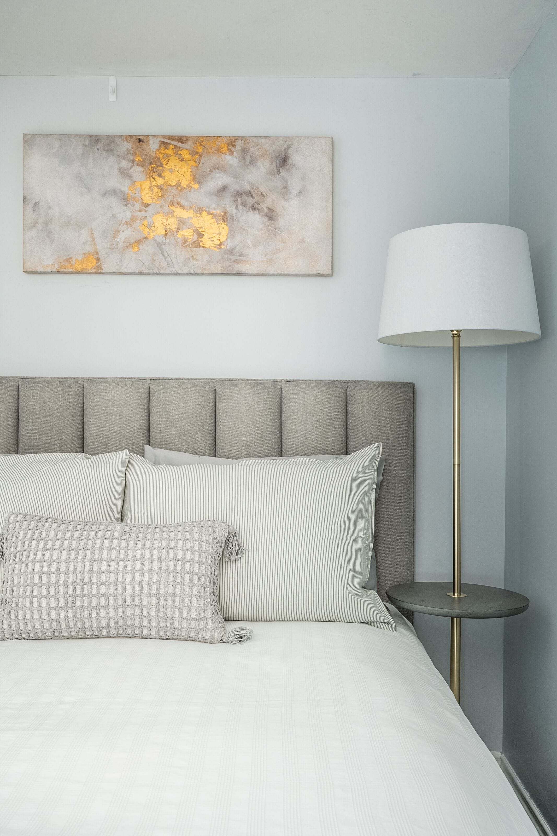 Una cama bien hecha con cabecera gris, una lámpara de noche y un cuadro abstracto colgado en la pared.