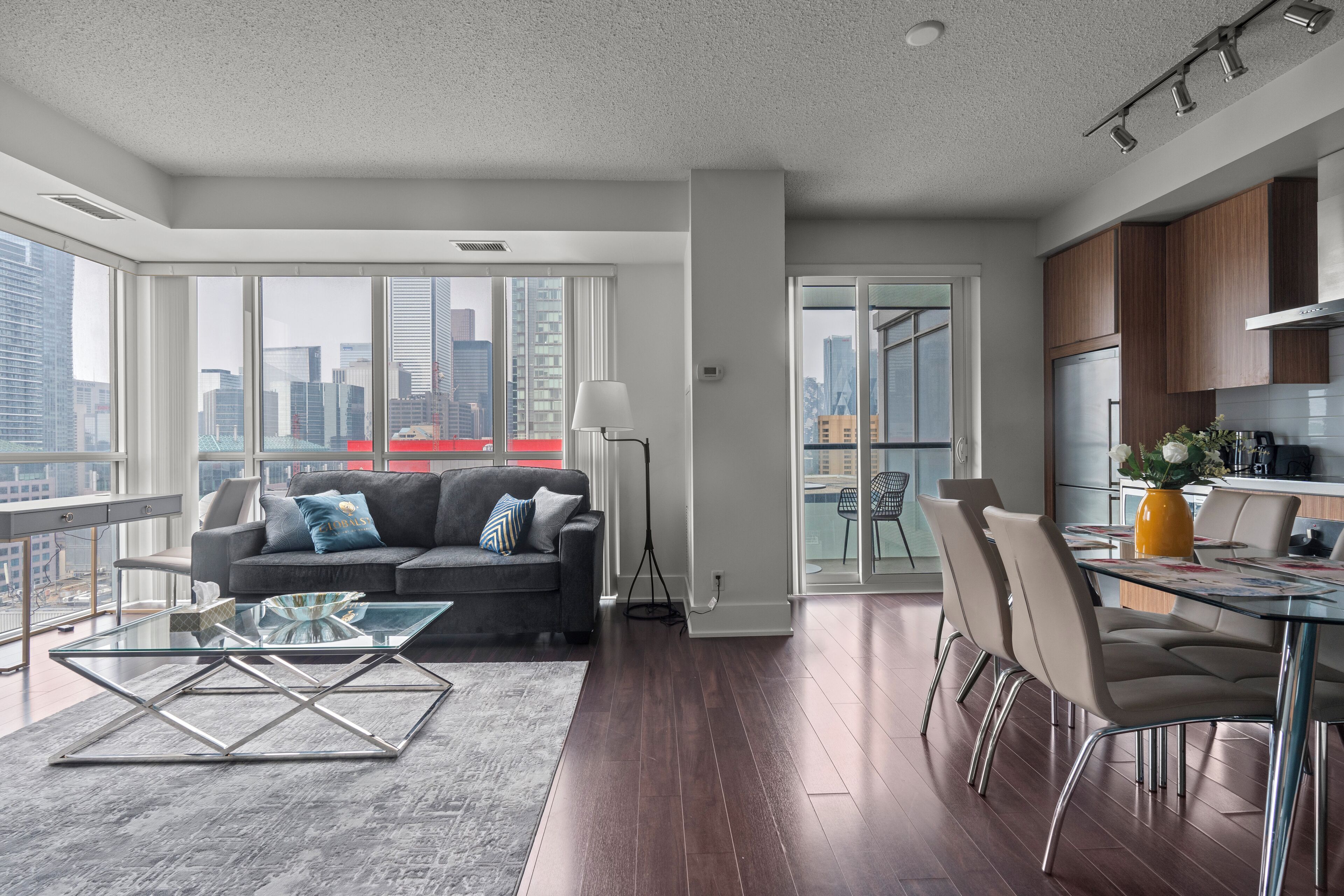 A modern apartment with a glass dining table, a sofa with cushions, a glass coffee table, and a city view through large windows.