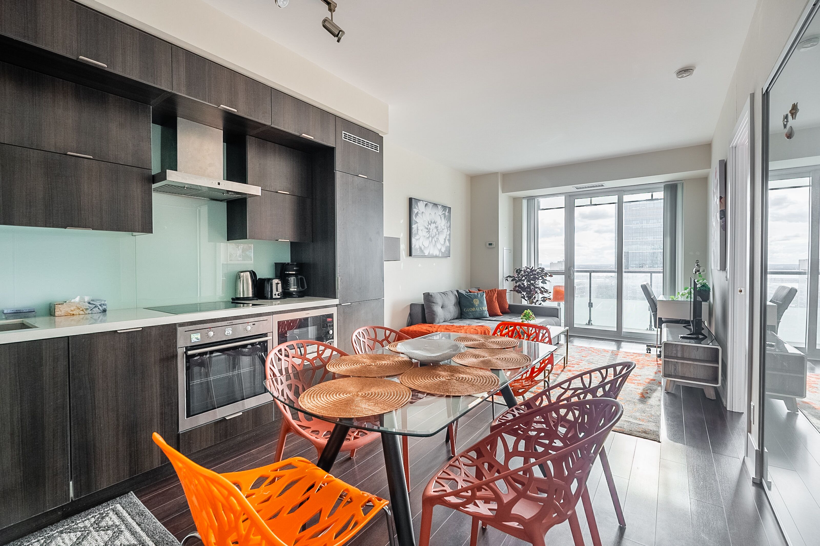 Una cocina moderna con armarios de madera oscura, una mesa de comedor de vidrio con sillas naranjas y un ventanal con vistas a un paisaje urbano.