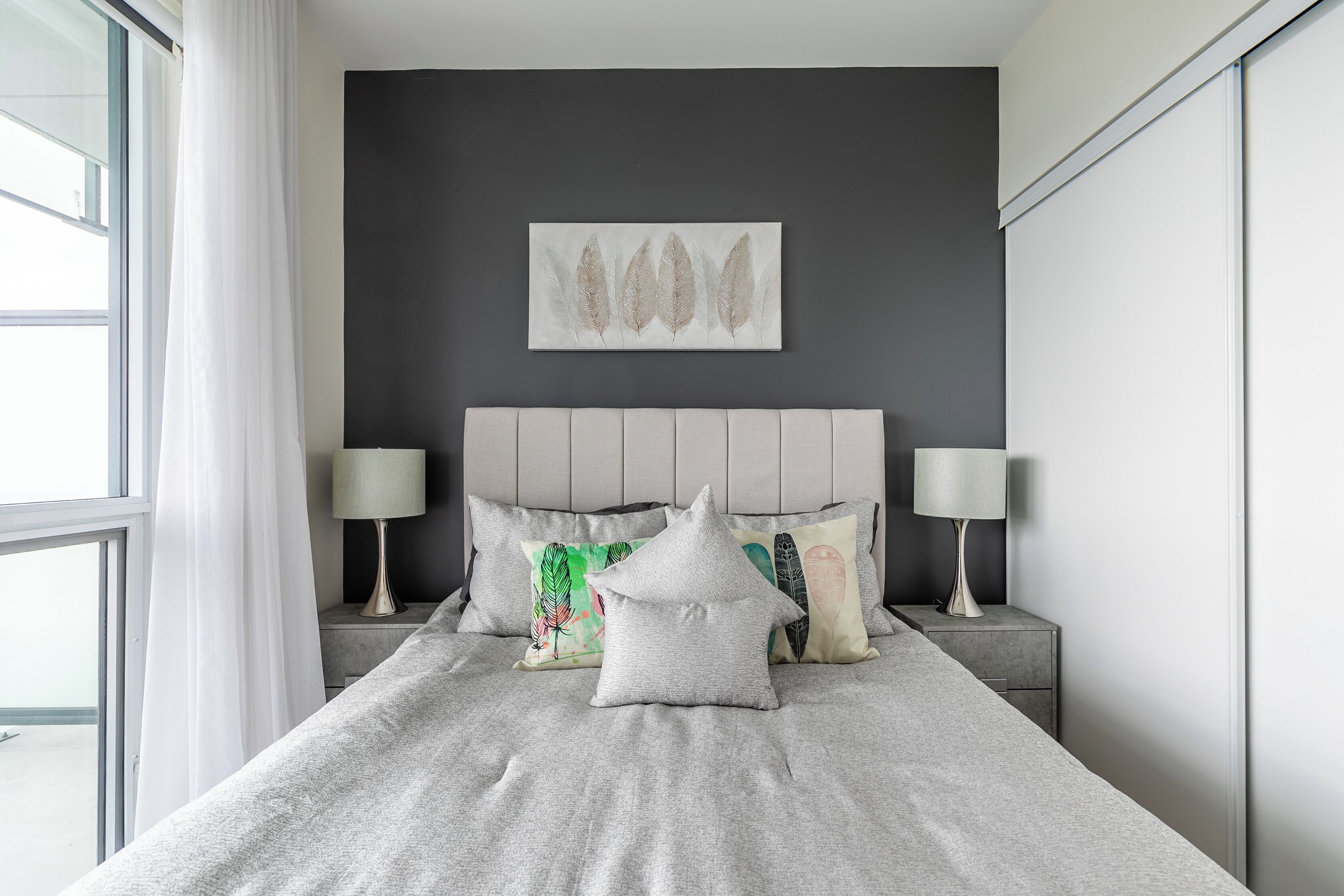 A modern bedroom with a grey bedspread, decorative pillows, a grey headboard, and a wall-mounted artwork.