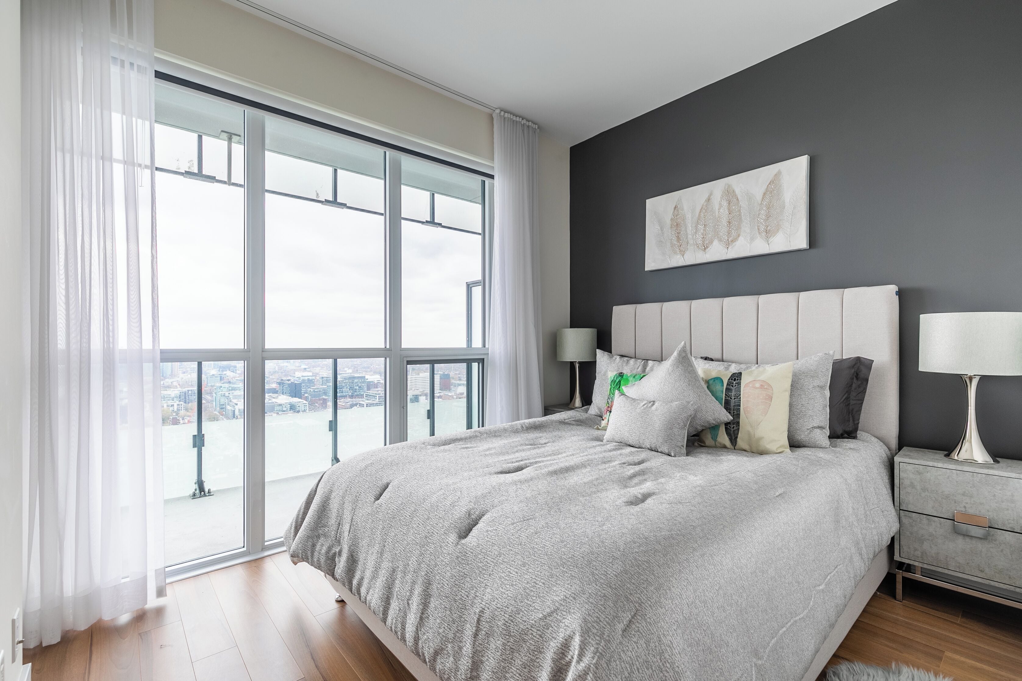 Un dormitorio moderno con un ventanal grande, una cama gris con muchos almohadones y una mesita de noche gris con una lámpara.