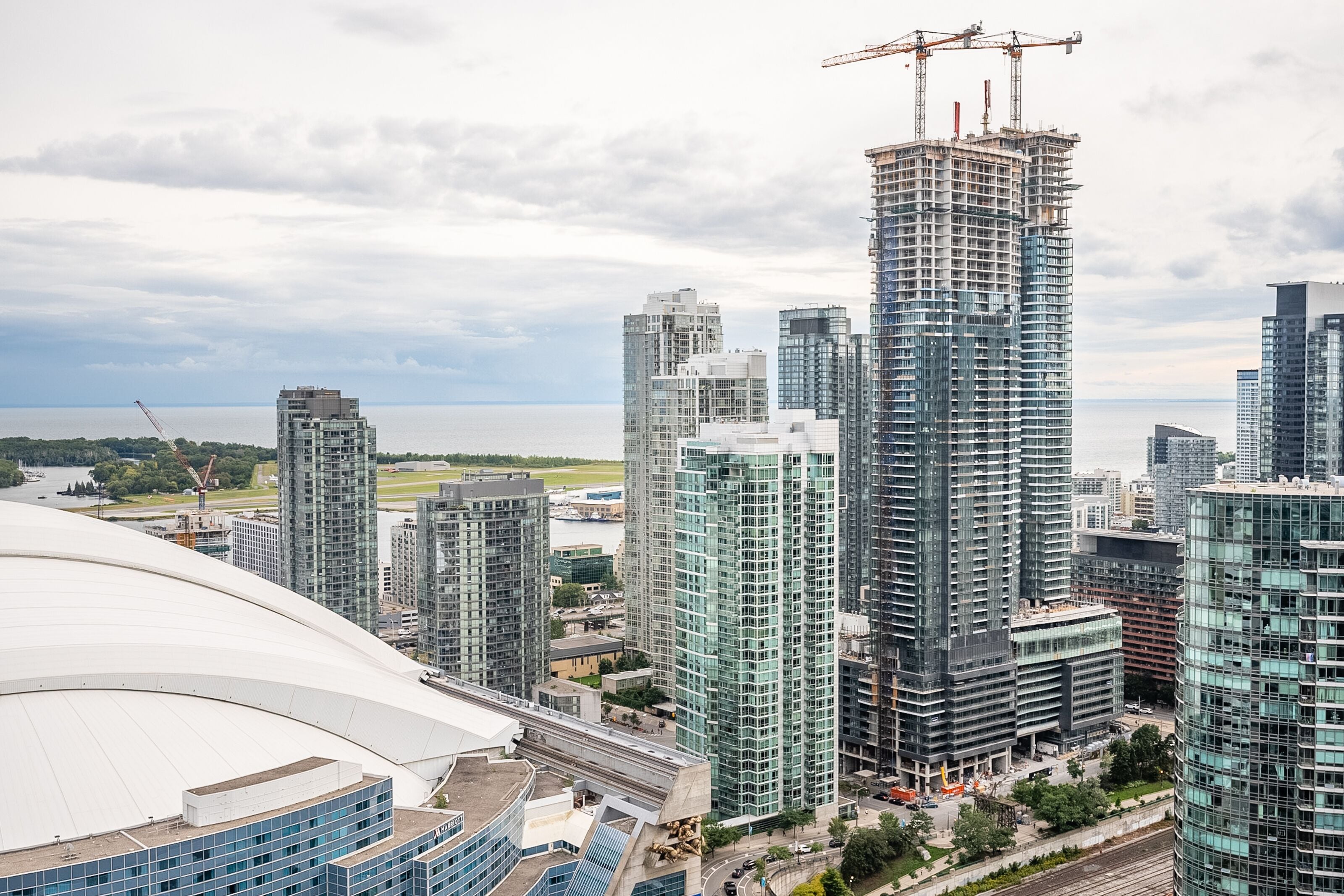 A high-rise building under construction with a crane, surrounded by other tall buildings and a stadium.