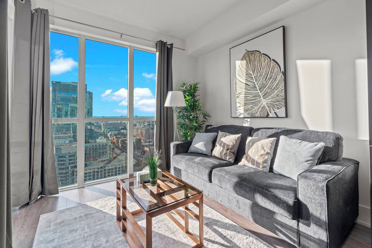 A modern living room with a grey sofa, a glass coffee table, and a large window offering a city view.