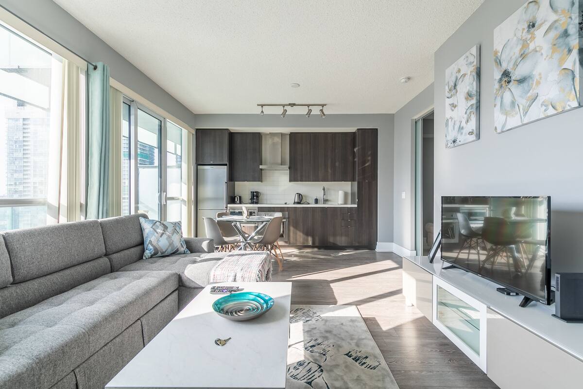 A modern living room with a grey sofa, a white coffee table, and a TV. There is a kitchen area with dark cabinets and a dining table.