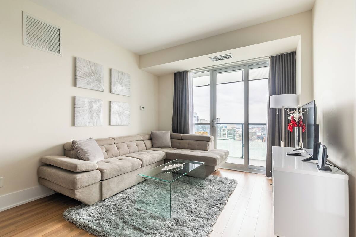 A modern living room with a beige sectional sofa, a glass coffee table, and a view of a cityscape through large windows.