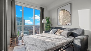 A modern bedroom with a large bed, a city view, a plant, and a framed artwork on the wall. - Modern Downtown Apartments (Toronto)
