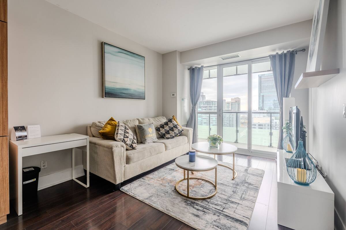 A modern living room with a sofa, a desk, a round coffee table, and a view of the city through large windows.